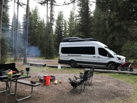 RV rental camper van at a smoky forest campsite. Picnic table cooking, active campfire, and chairs set up for relaxation.