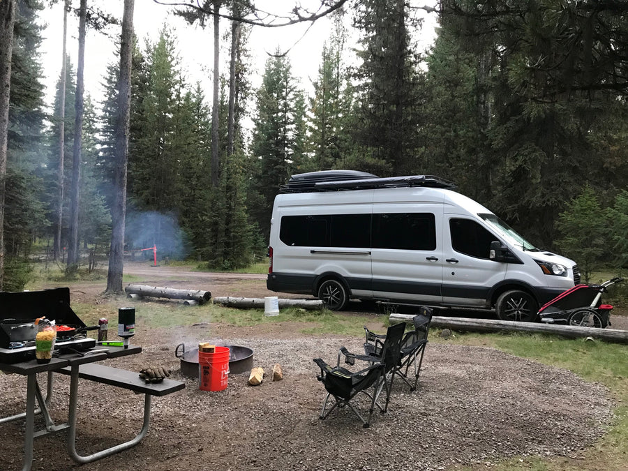 RV rental camper van at a smoky forest campsite. Picnic table cooking, active campfire, and chairs set up for relaxation.