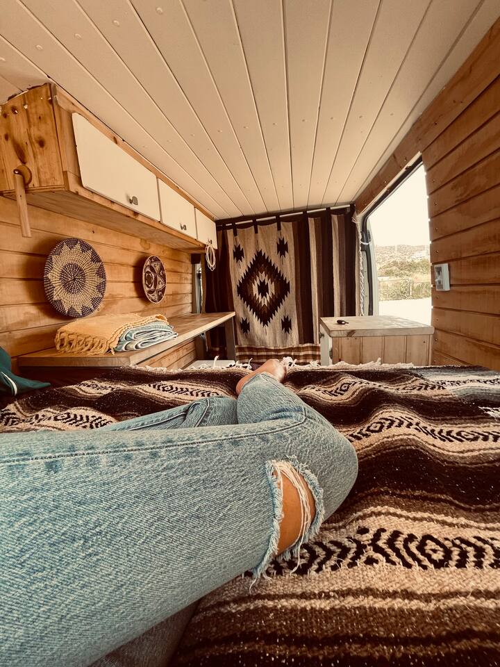 Cozy RV rental interior: rustic wood, decorative baskets, patterned blanket, & relaxing legs. Van life with open views. — photo 11