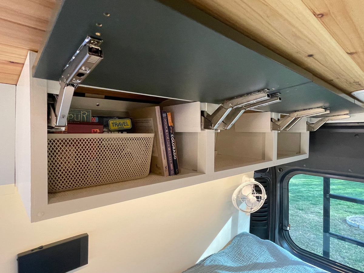 RV rental overhead storage: books, travel games, and a basket in open cabinets, enhancing comfort with a fan & window view. — photo 19
