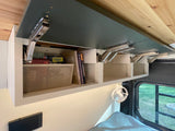 RV rental overhead storage: books, travel games, and a basket in open cabinets, enhancing comfort with a fan & window view.