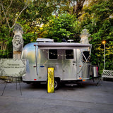 Airstream rv rental transformed into a 'Versailles Salon de Beauté' with awning, lights, & glowing display in a lush garden.