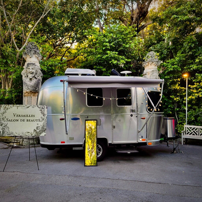 Airstream rv rental transformed into a 'Versailles Salon de Beauté' with awning, lights, & glowing display in a lush garden.