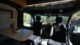 Inside this rv rental: front seats, dash, and a dining table with fruit & lamp. Scenic trees visible through the windshield.