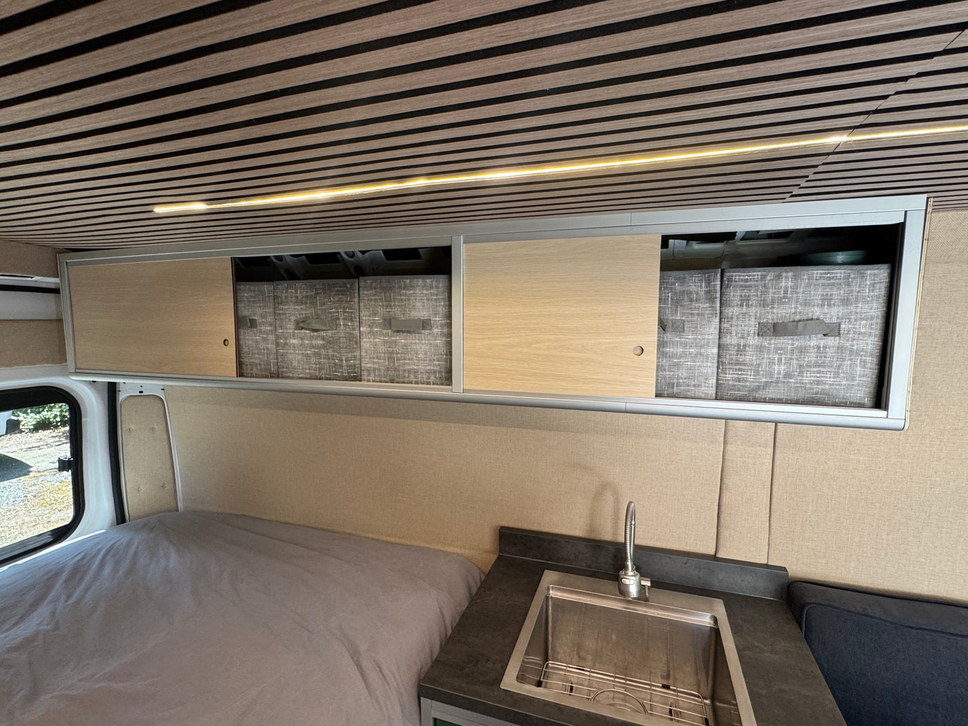 Spacious RV rental interior with bed, kitchen sink, overhead storage, and modern slat ceiling with lighting.