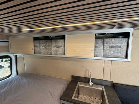 Spacious RV rental interior with bed, kitchen sink, overhead storage, and modern slat ceiling with lighting.