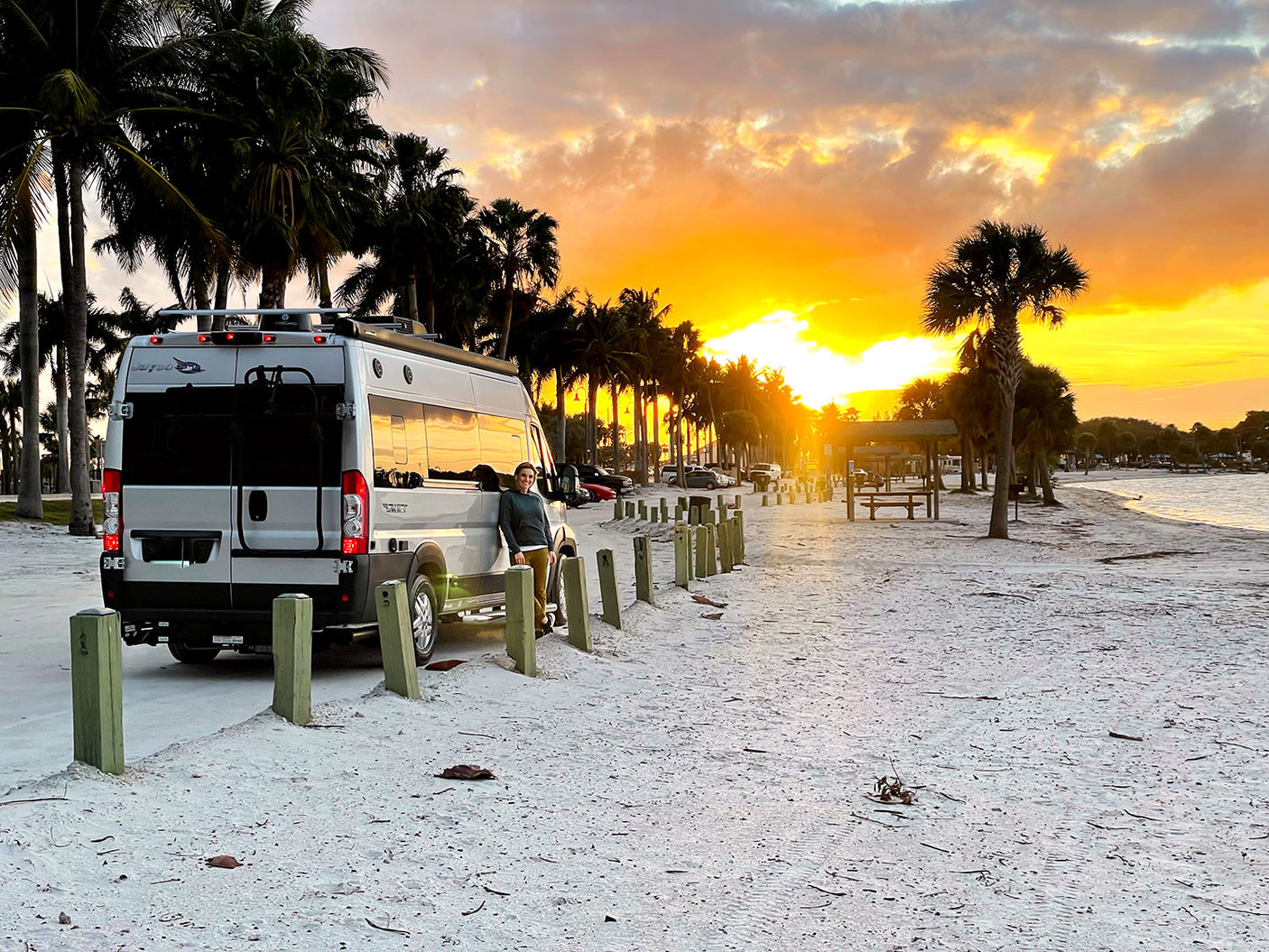 Golden hour glow: An RV rental parked on a sandy beach, person enjoys a stunning palm tree sunset.