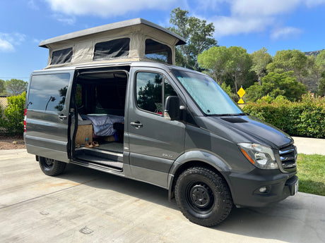 A grey Mercedes Sprinter pop-top camper van is parked with its side door open, revealing the interior. Ideal RV rental for travel.
