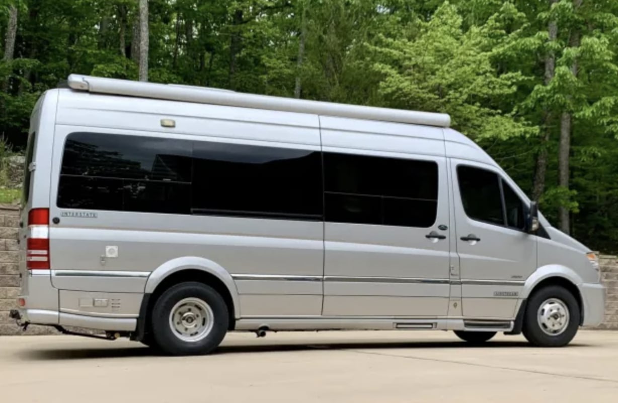 A silver Airstream Interstate camper van, perfect for RV rental travel. This sleek, modern RV is parked outdoors with trees.