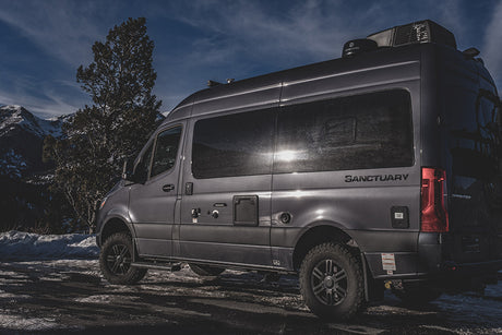 A gray Sprinter van RV rental, branded 'Sanctuary', is parked in a snowy mountain setting, ready for your next adventure.
