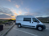 White camper van (RV rental) parked at sunset by the ocean, with distant hills & a picnic table.