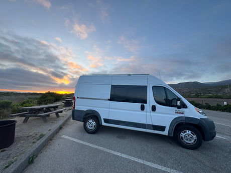 White camper van (RV rental) parked at sunset by the ocean, with distant hills & a picnic table.
