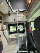 Compact RV rental interior: Silver ladder to pop-top bed, light seating area with table, and campground view.