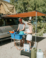 RV rental trip: A couple washes dishes outdoors using a portable HOTTAP and sink setup next to their vehicle with a rooftop tent.