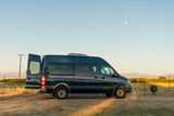Explore nature with this rv rental! Dark blue campervan parked in a grassy field with open rear door under a pale moon, mountains beyond.