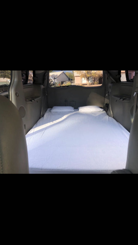 Minivan converted for travel, ideal for RV rental. Rear interior shows a bed with white sheets & pillows for car camping.