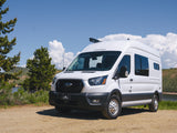 White Ford Transit campervan RV rental on a dirt road, by a scenic lake with trees and hills under a blue sky.