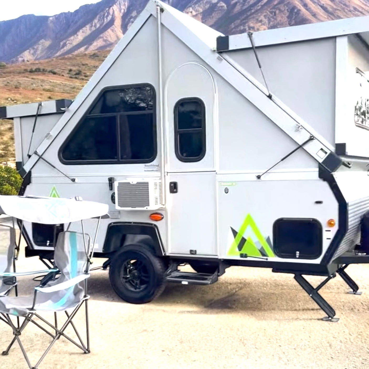 An A-frame RV rental is set up in a mountainous landscape with camping chairs. Perfect for outdoor adventures.