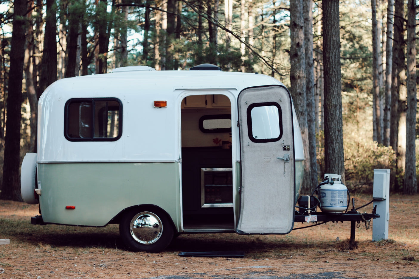 Charming white & green rv rental awaits in a peaceful forest. Its open door reveals an inviting space for camping adventures.
