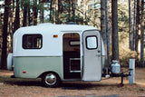 Charming white & green rv rental awaits in a peaceful forest. Its open door reveals an inviting space for camping adventures.