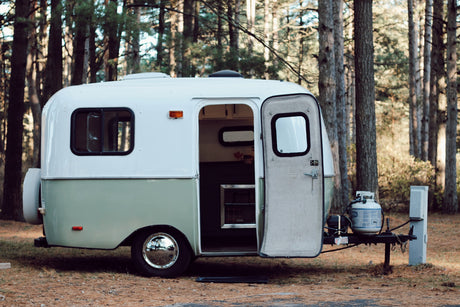 Charming white & green rv rental awaits in a peaceful forest. Its open door reveals an inviting space for camping adventures.