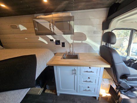 This RV rental interior features a compact kitchen with a sink, overhead cabinets, and a cozy bed on the left, perfect for travel.