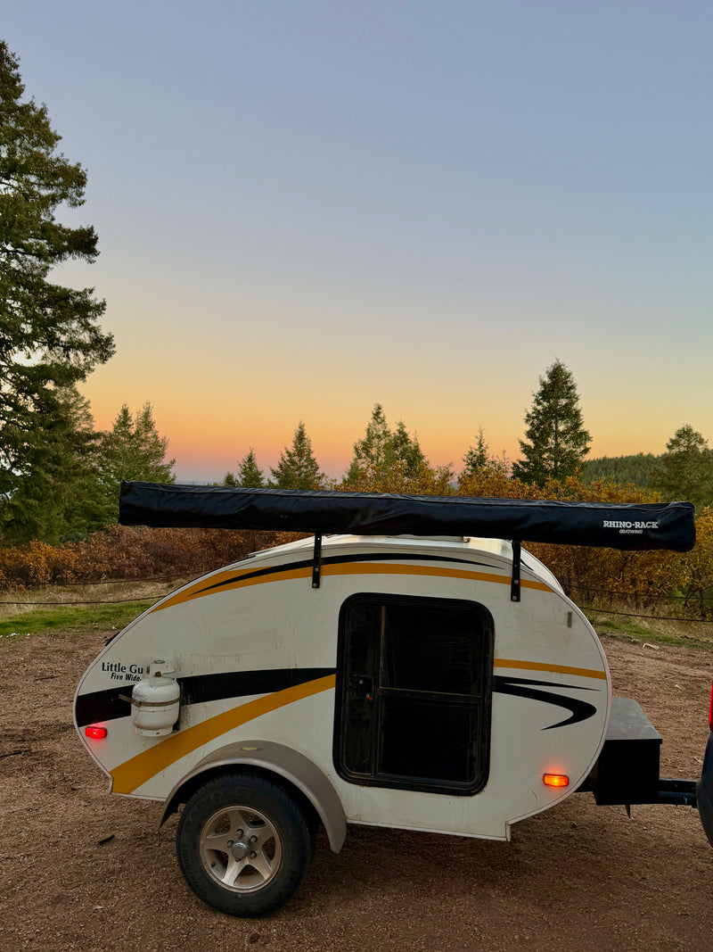 A white teardrop RV rental camper with a black awning under a golden sunset sky, surrounded by pine trees. Ready for camping. — photo 3