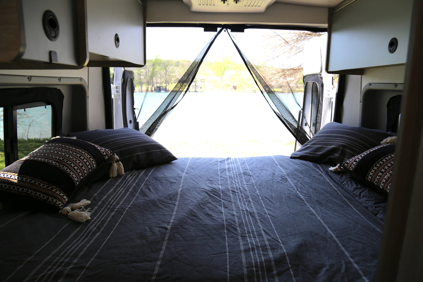 Comfortable RV rental bed with scenic lake view from open rear doors, featuring a mesh screen for peaceful travel.