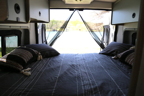 Comfortable RV rental bed with scenic lake view from open rear doors, featuring a mesh screen for peaceful travel.