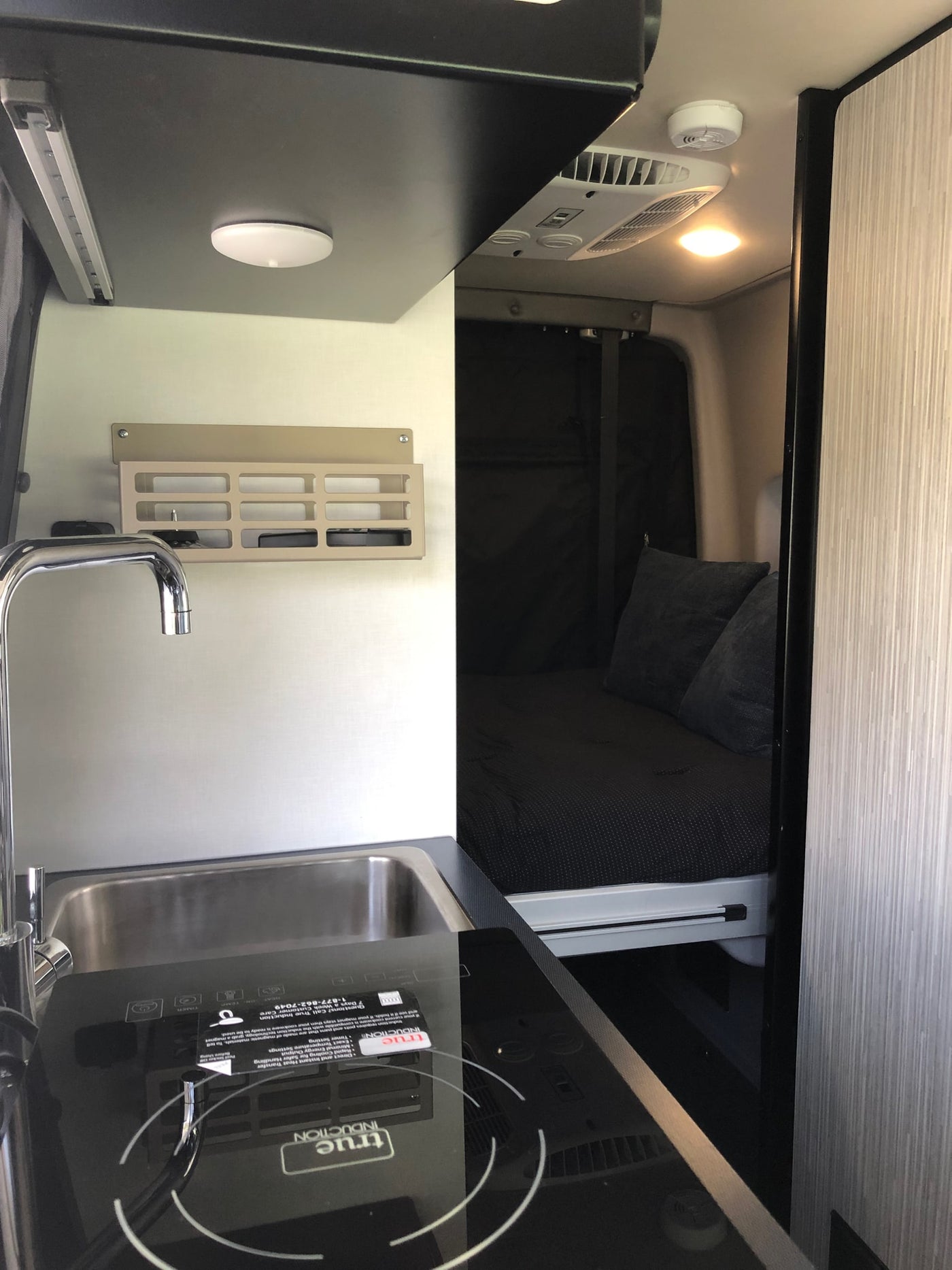 This RV rental shows a compact interior with a modern kitchen, featuring a sink and True Induction cooktop, alongside a comfy sleeping area. — photo 13