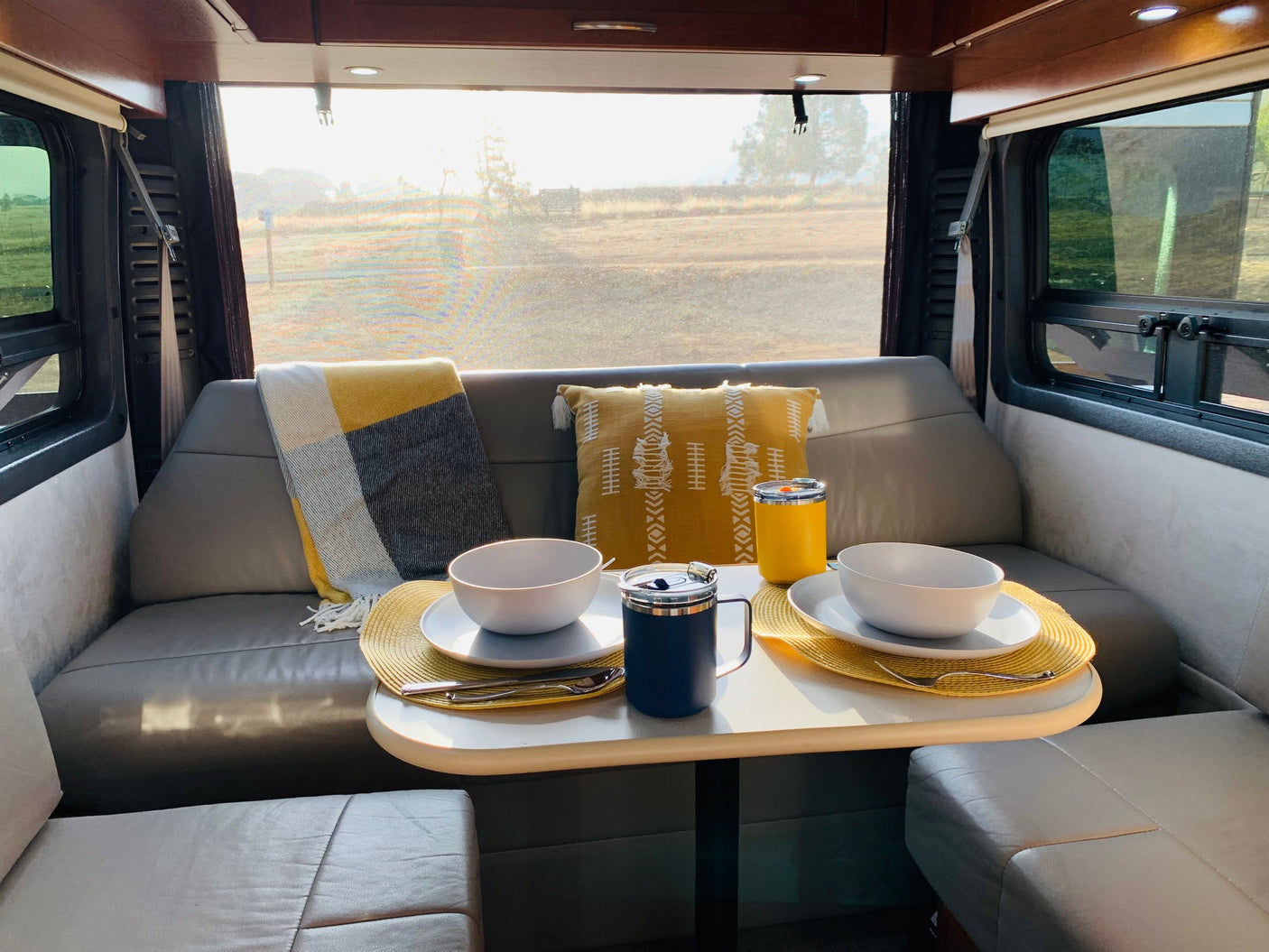 Bright RV rental interior: a cozy dining area set for two with white bowls, yellow placemats, and large scenic windows.