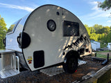Compact white teardrop RV rental shown outdoors. This travel trailer features mountain graphics and is ready for camping.