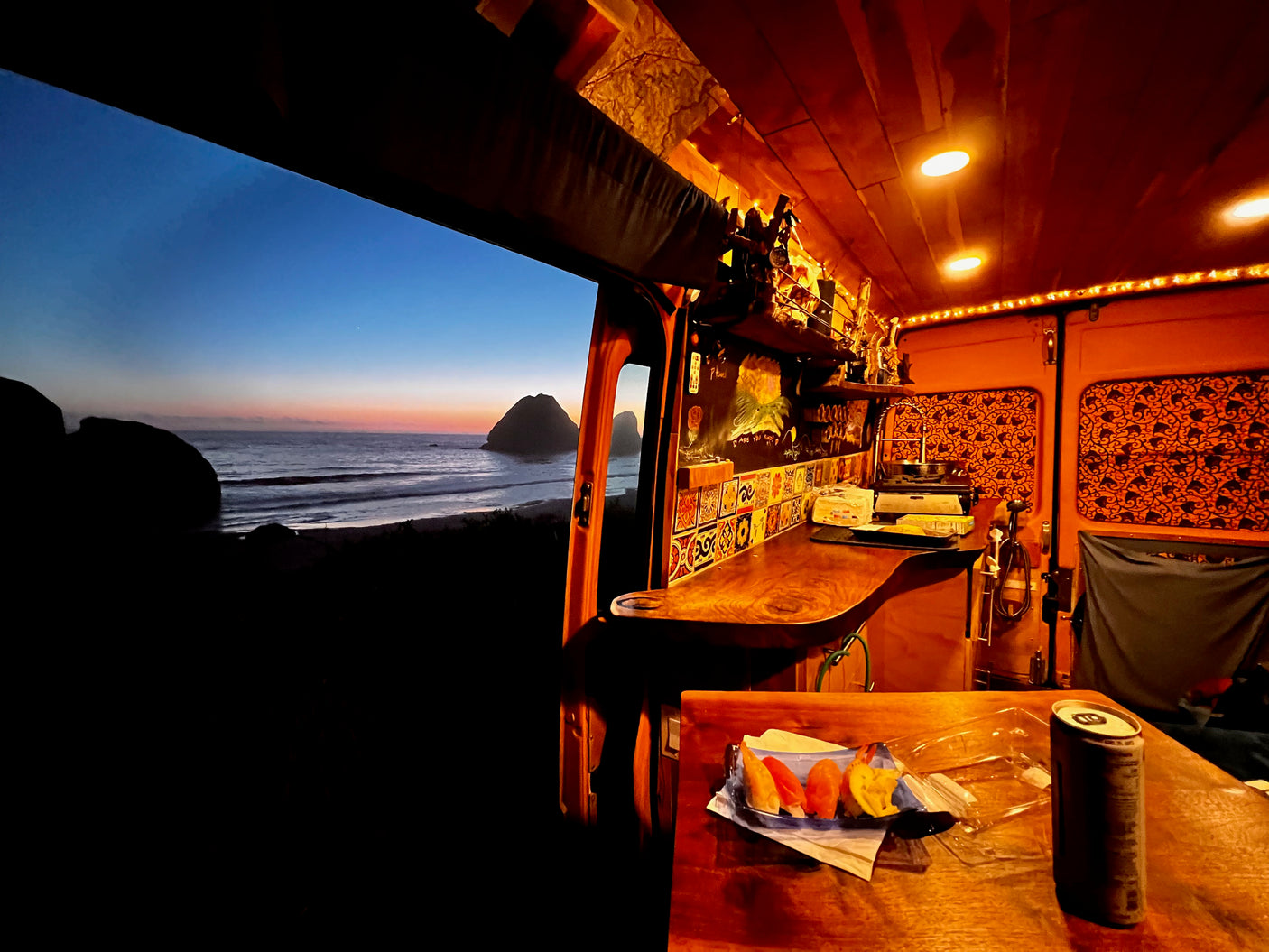 Cozy RV rental interior with ocean sunset view. Warm lights illuminate the counter and a table with food inside.