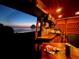 Cozy RV rental interior with ocean sunset view. Warm lights illuminate the counter and a table with food inside.