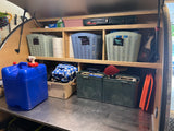 Inside an RV rental: shelves filled with organized camping gear. Labeled baskets hold 'Yard Toys,' 'Breads,' 'Breakfast,' next to a stove & water.