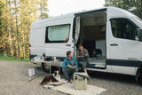 Enjoying an RV rental adventure: A couple and their dogs relax, cooking and dining outdoors next to their camper van.