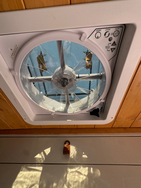 Inside an RV rental, a MaxxAir ceiling fan provides ventilation. Its control panel and mesh screen are visible, with sky above.