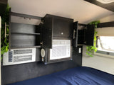 Modern RV rental bedroom featuring dark cabinetry, two integrated AC units, accent lighting, and green foliage above a blue bed.