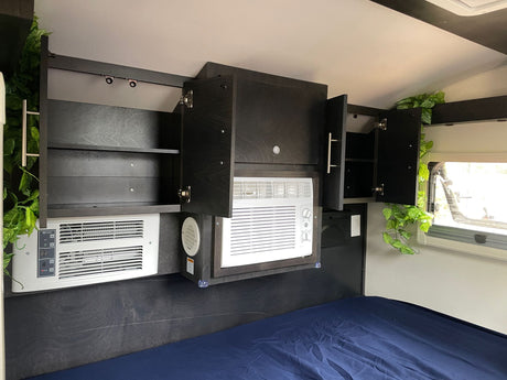Modern RV rental bedroom featuring dark cabinetry, two integrated AC units, accent lighting, and green foliage above a blue bed.
