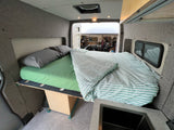 Cozy RV rental interior with a comfy bed, striped duvet, compact sink, & view out the open rear doors.