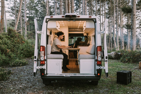 This RV rental offers a cozy interior setup for remote work. A man is on his laptop, enjoying nature from the back of the camper van.