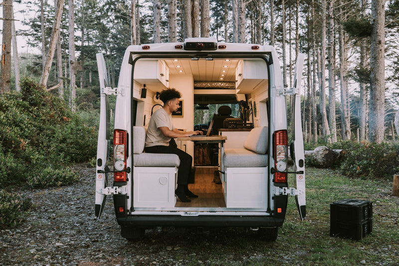 This RV rental offers a cozy interior setup for remote work. A man is on his laptop, enjoying nature from the back of the camper van. — photo 14