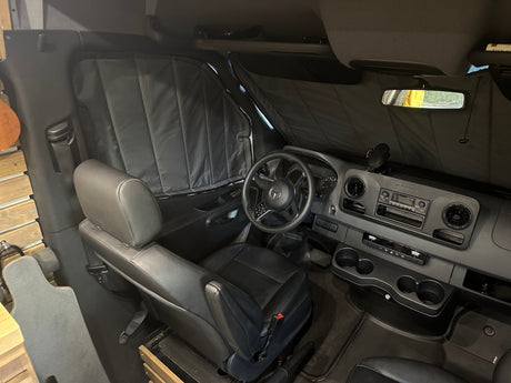 Inside this RV rental: driver's cabin with Mercedes dashboard & insulated privacy shades. A longboard hints at adventure.