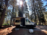 A white camper van rv rental with an extended awning is parked at a sunlit forest campsite, ready for outdoor living.