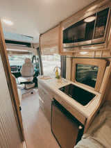 Inside this RV rental: front seating, a compact kitchen with microwave, fridge, and cooktop, plus a cozy sleeping area.