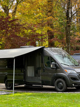Dark grey RV rental camper van with open door and extended awning, parked among vibrant autumn trees.