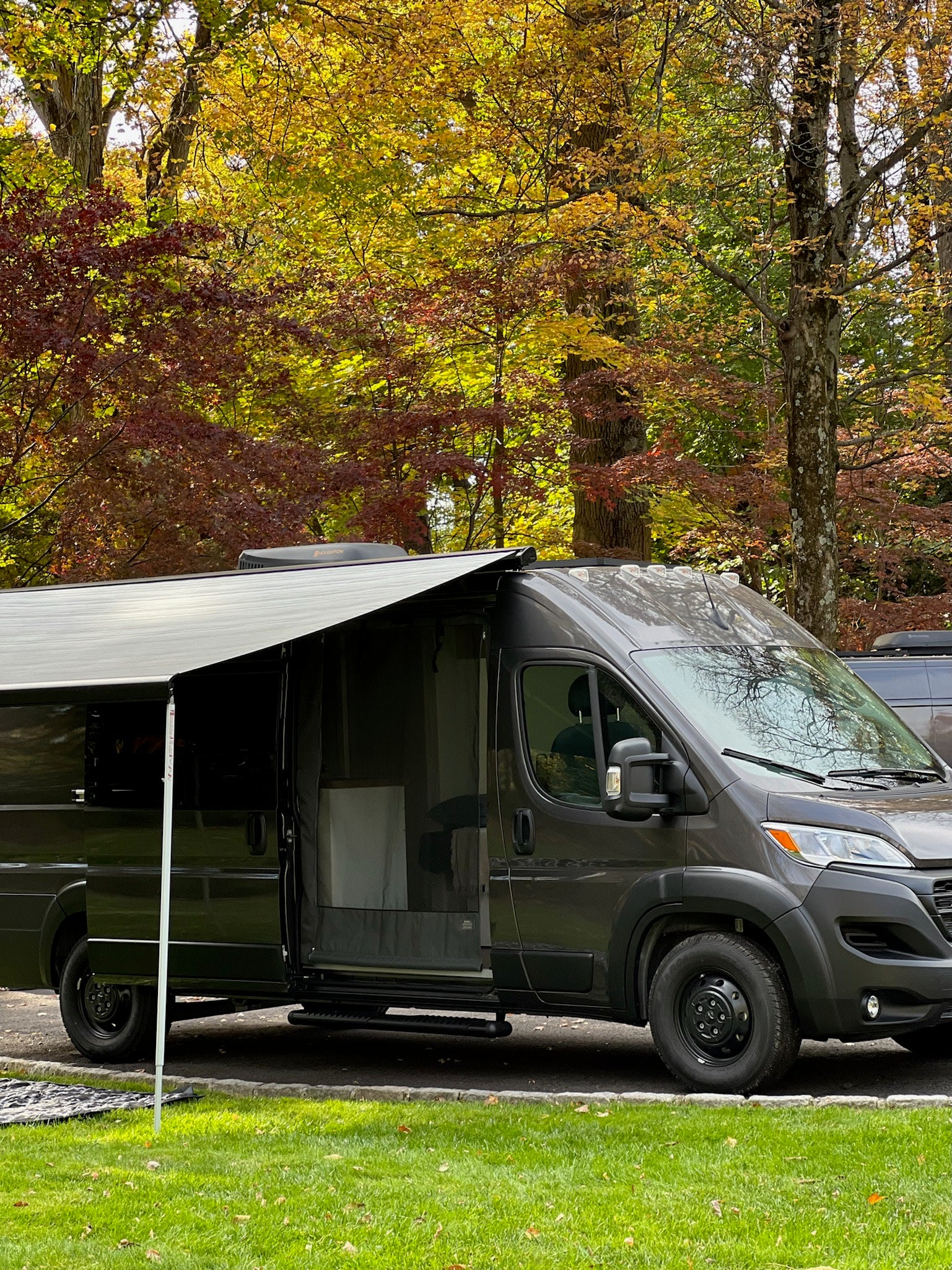 Dark grey RV rental camper van with open door and extended awning, parked among vibrant autumn trees.