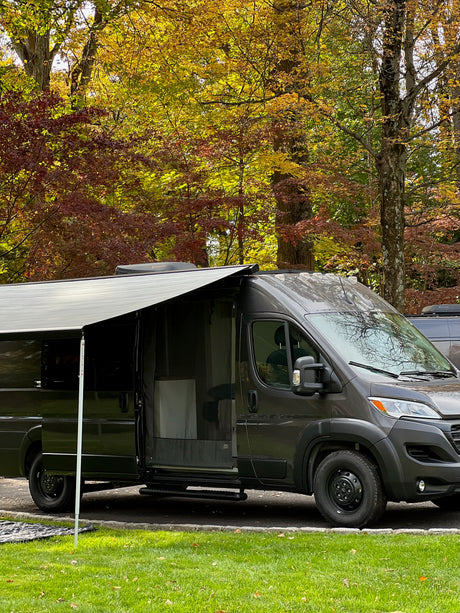 Dark grey RV rental camper van with open door and extended awning, parked among vibrant autumn trees.