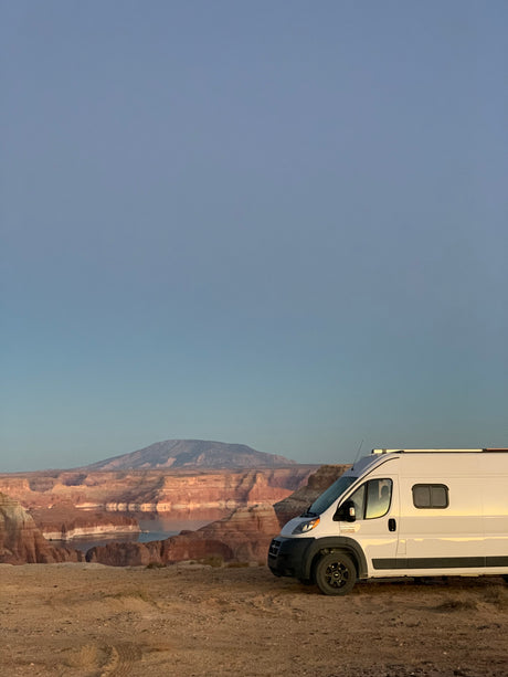 This white RV rental enjoys a breathtaking view of majestic red cliffs and a calm lake from a rugged dirt road.
