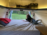 Inside an rv rental van: A child lounges on the bed, admiring nature through the open back, with overhead storage.